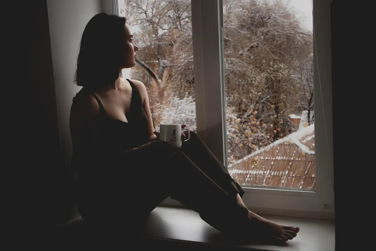 Woman sitting alone by a window, looking out at winter trees